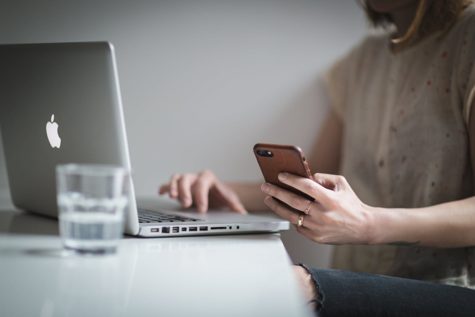 Person using a laptop and smartphone at a table with a glass of water.