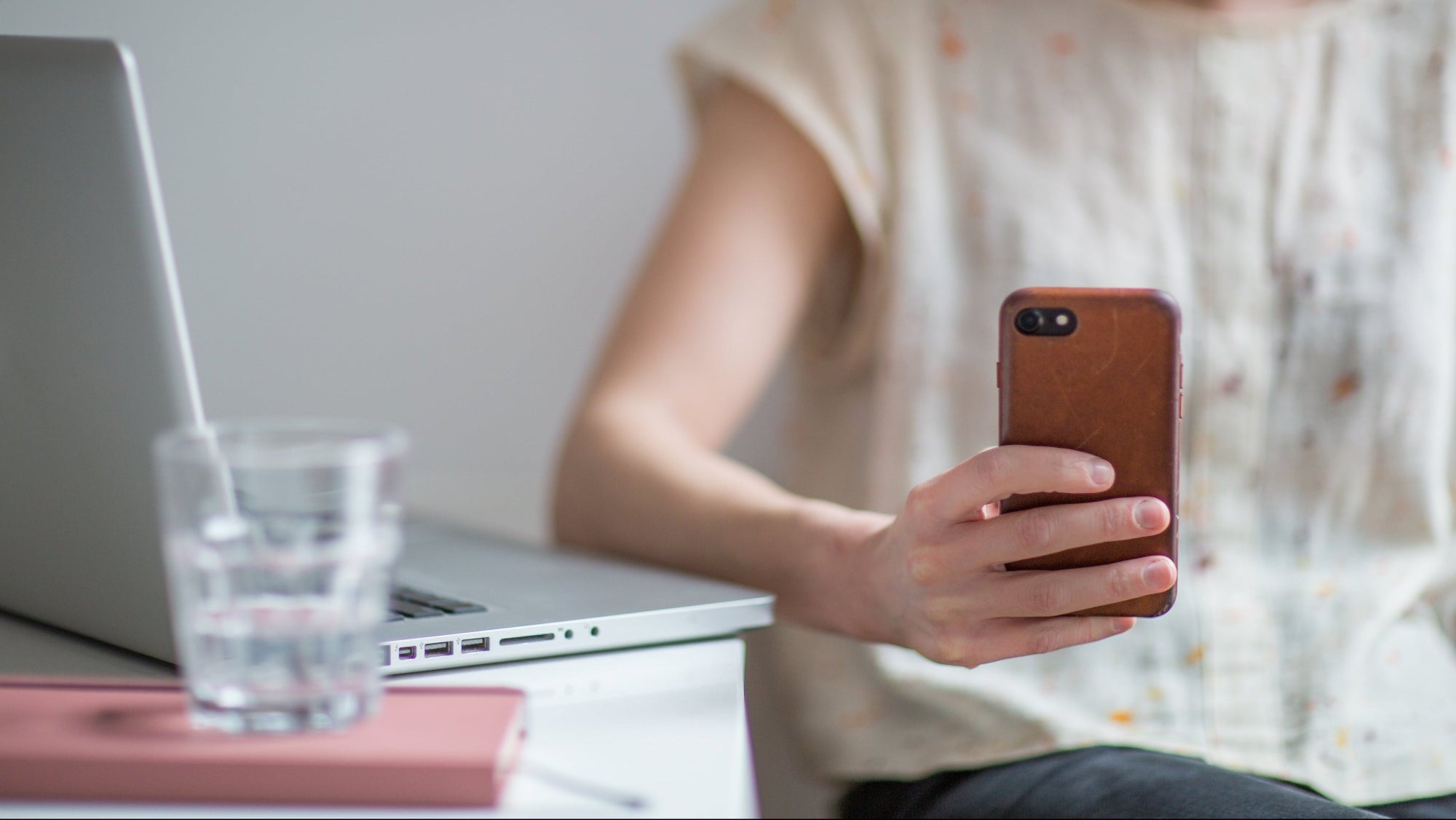 Person holding a smartphone with a laptop and glass of water on a table